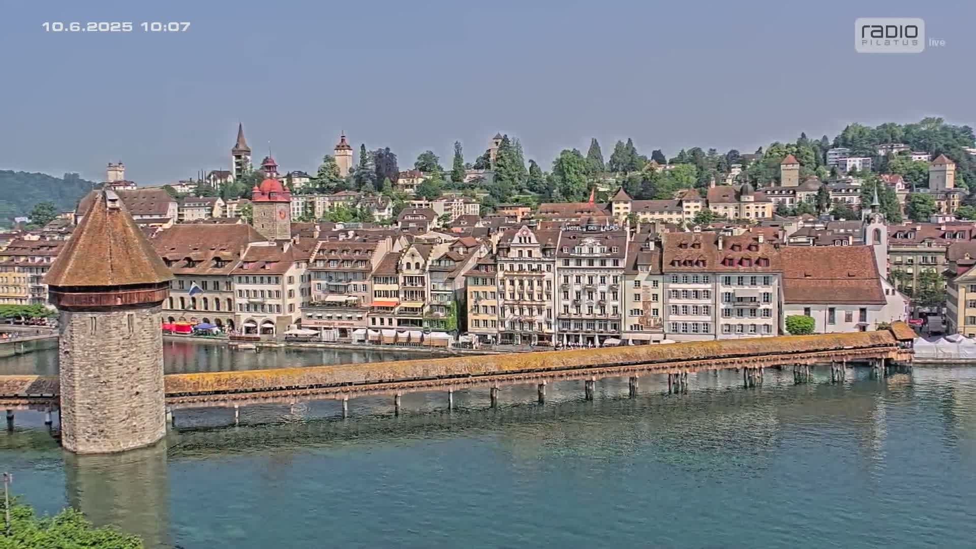 A covered wooden bridge spans a body of water in front of a city with numerous buildings, under a clear sky.