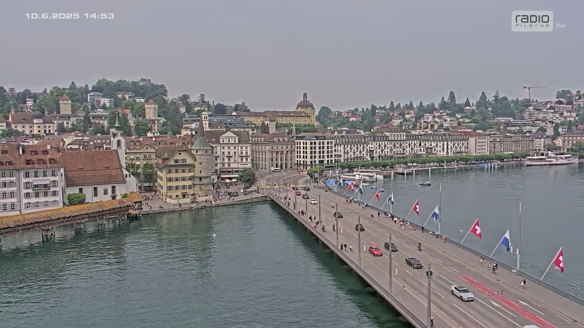 An aerial view shows a bridge over water with vehicles and pedestrians, leading to a city with numerous buildings under an overcast sky.