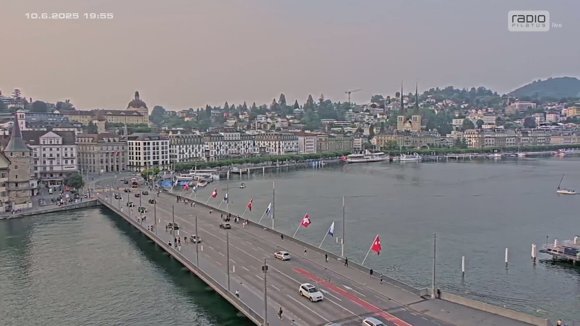 A long bridge with cars and pedestrians crosses a body of water toward a city with many buildings under a hazy, dusky sky.