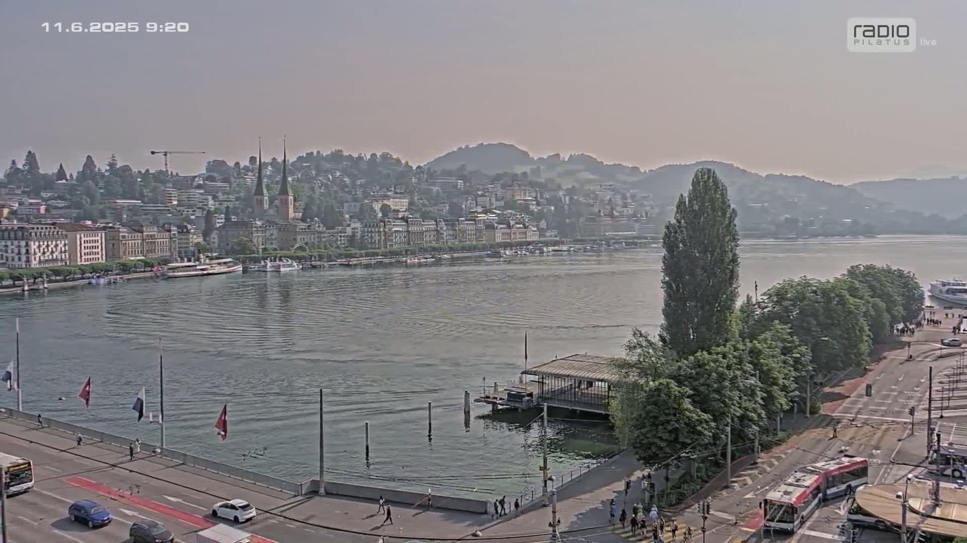 A hazy view of a city on a lake, with boats moored along the waterfront and a roadway with buses and cars visible in the foreground.