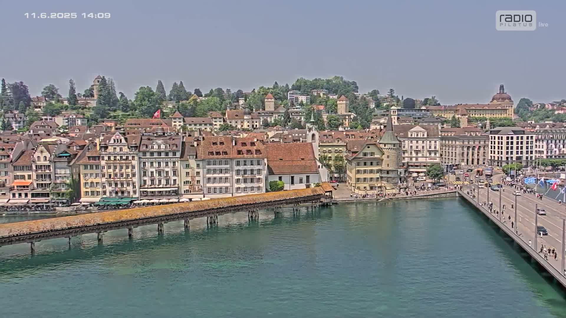 A picturesque waterfront town on a sunny day, featuring a long covered wooden bridge extending over a calm body of water, with numerous buildings lining the shore.