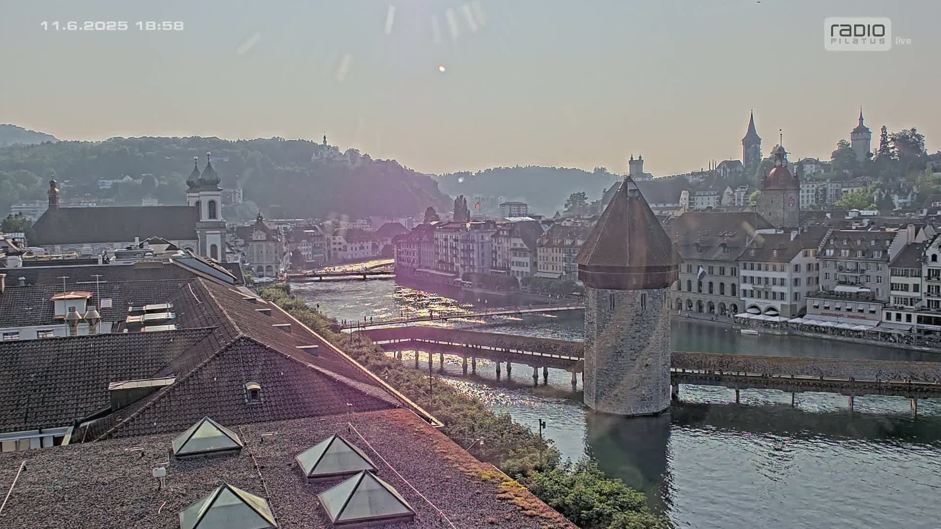 A hazy, sunlit view of a European city with a river running through it, showing several buildings, a covered bridge, and a water tower.