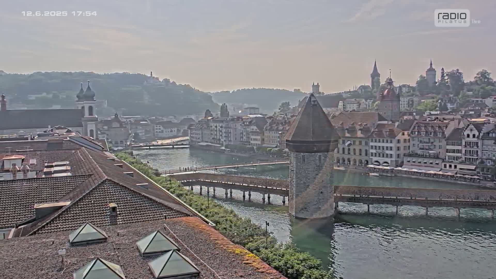 A hazy view of a European city with a river, a covered wooden bridge, and a stone tower, all under a bright, somewhat hazy sky.
