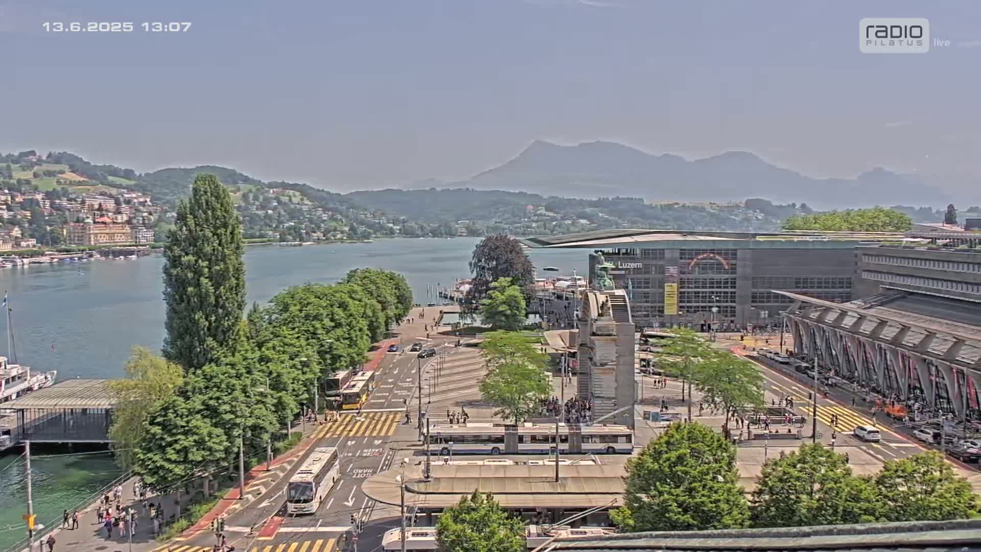 An elevated view of a city lakeside with clear skies, showing a large plaza with buses and pedestrians, a body of water, and mountains in the background.