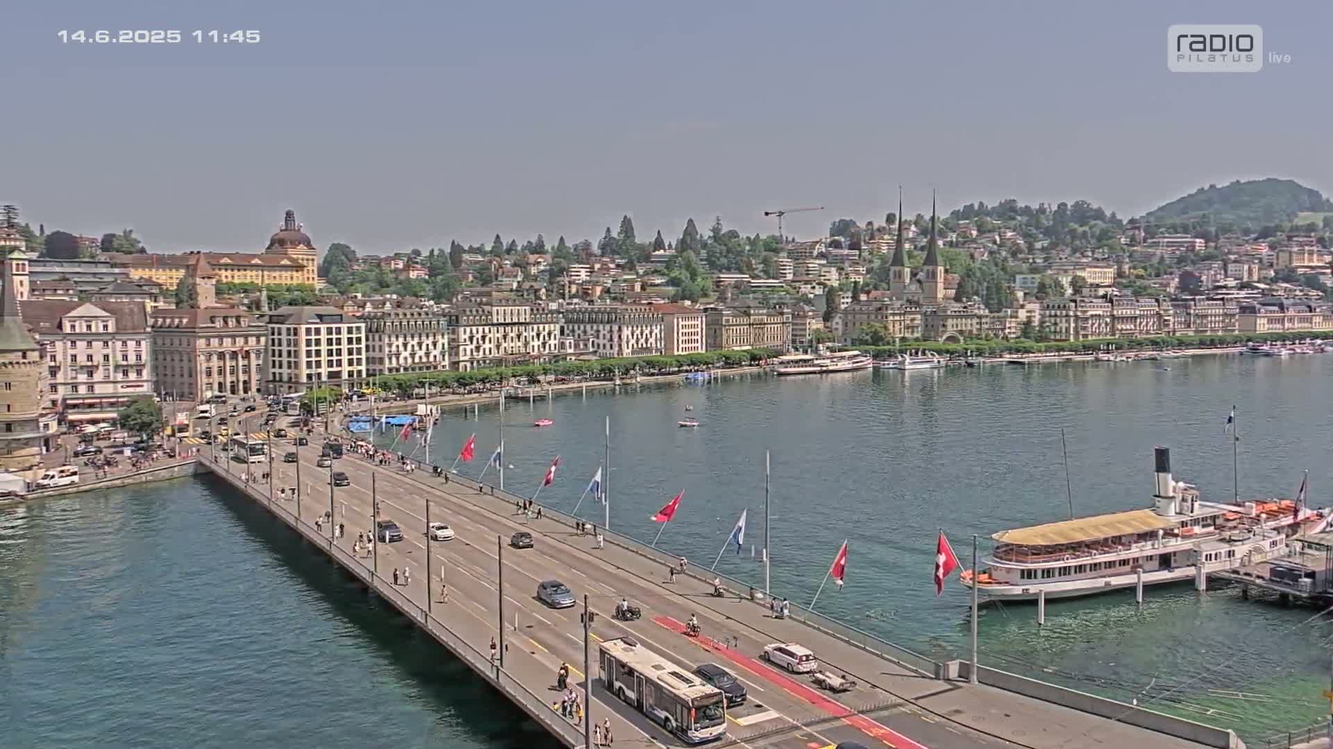A bridge with cars and pedestrians stretches across a calm lake toward a city of tall buildings under a clear, sunny sky.