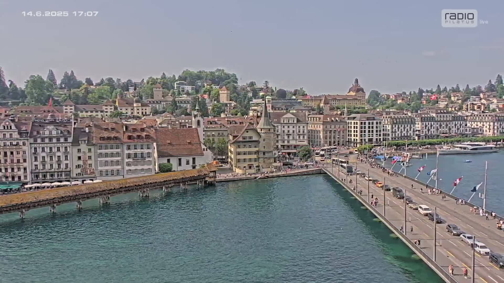 A sunny view of a city with numerous buildings along a body of water, a bridge with pedestrians and vehicles, and boats in the distance.