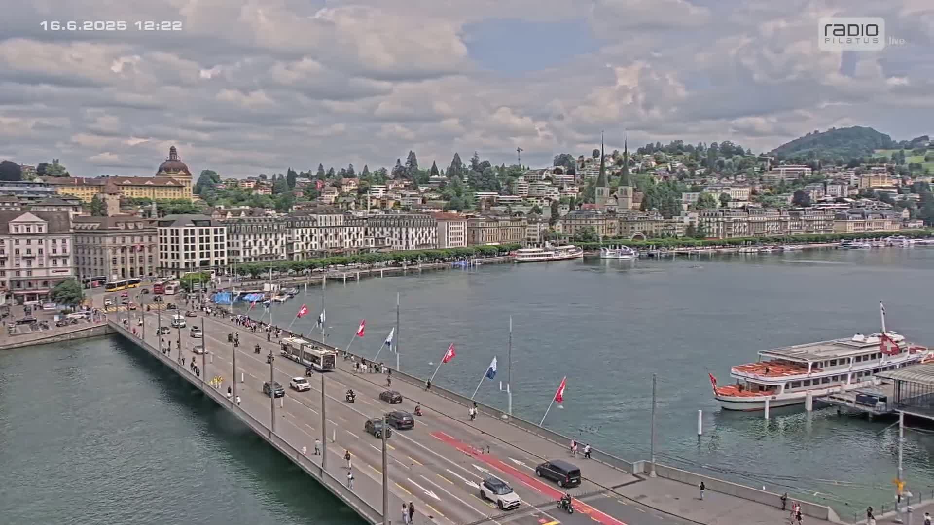 A partly cloudy day reveals a city waterfront with a bridge carrying traffic and pedestrians, boats on the water, and buildings rising on a hillside.