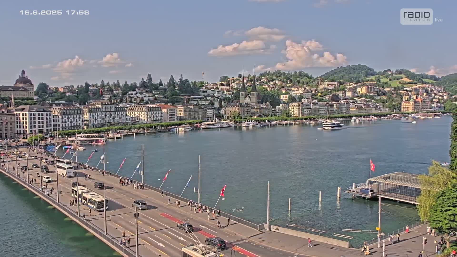 A bridge over a calm lake with a city and hills in the background under a partly cloudy sky.