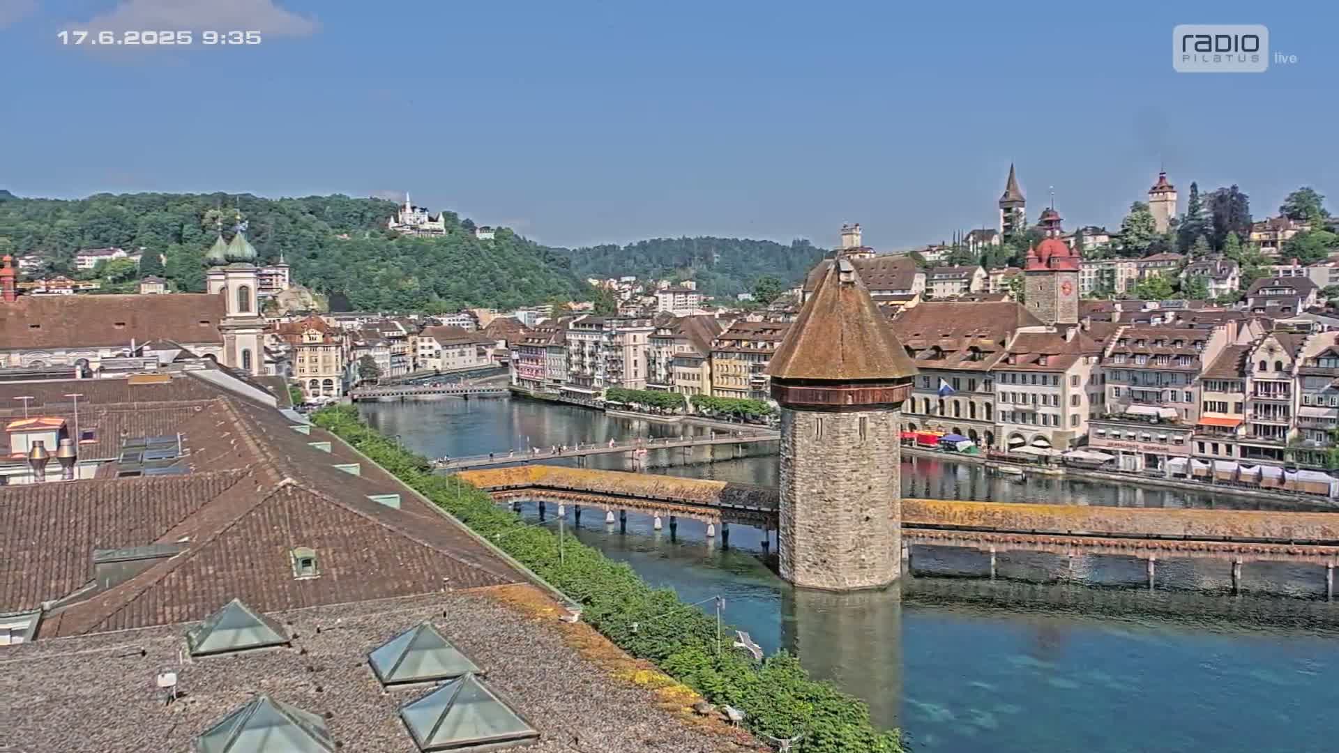 A picturesque European town with a river flowing through it, featuring a stone tower on a covered bridge, under a clear sunny sky.