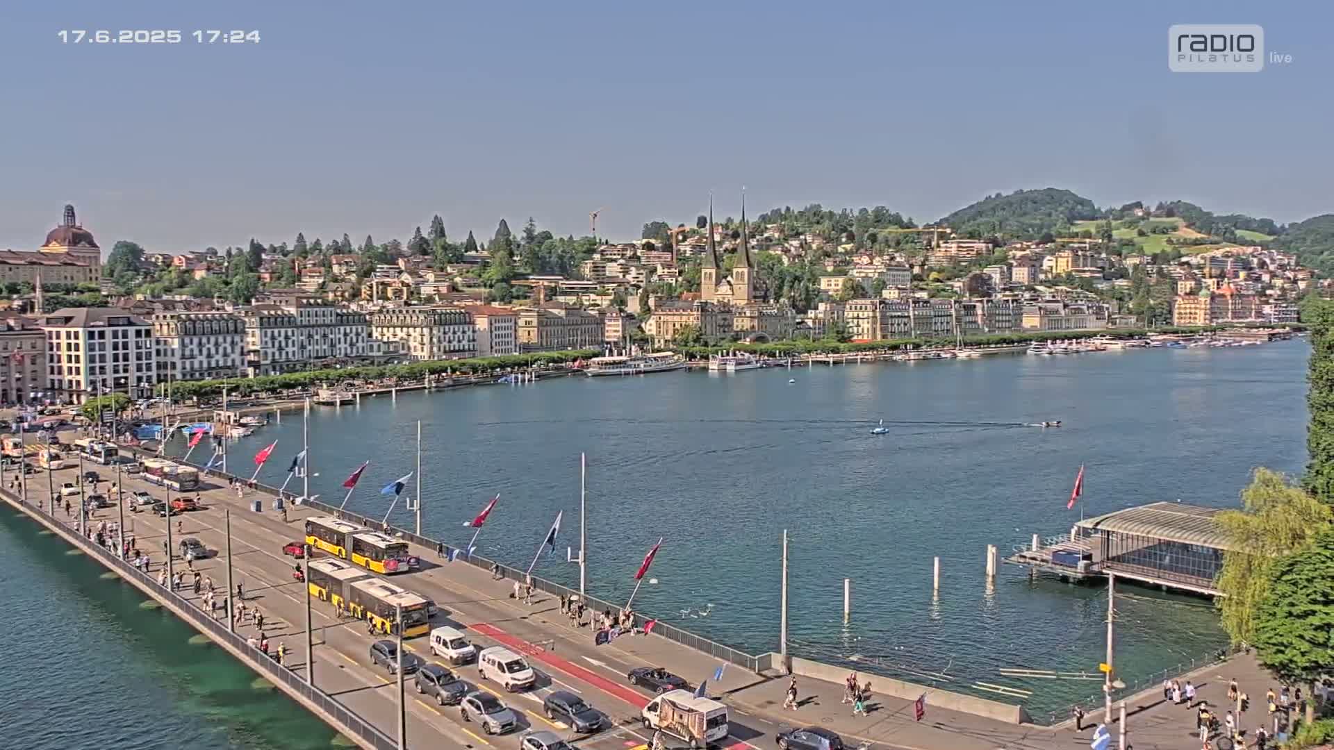 A bridge with traffic and pedestrians overlooks a calm lake, with a city of buildings and hills in the background on a sunny day.