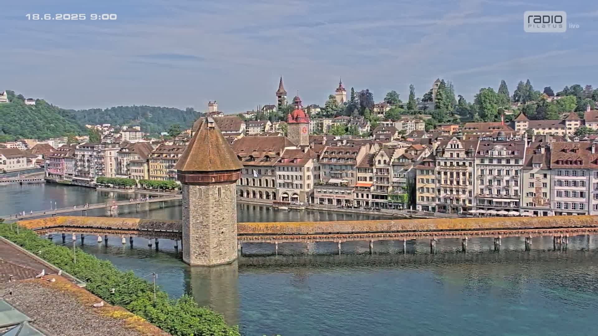 A stone tower stands at the end of a covered wooden bridge spanning a calm body of water, with a town of old buildings and lush green hills in the background on a sunny day.