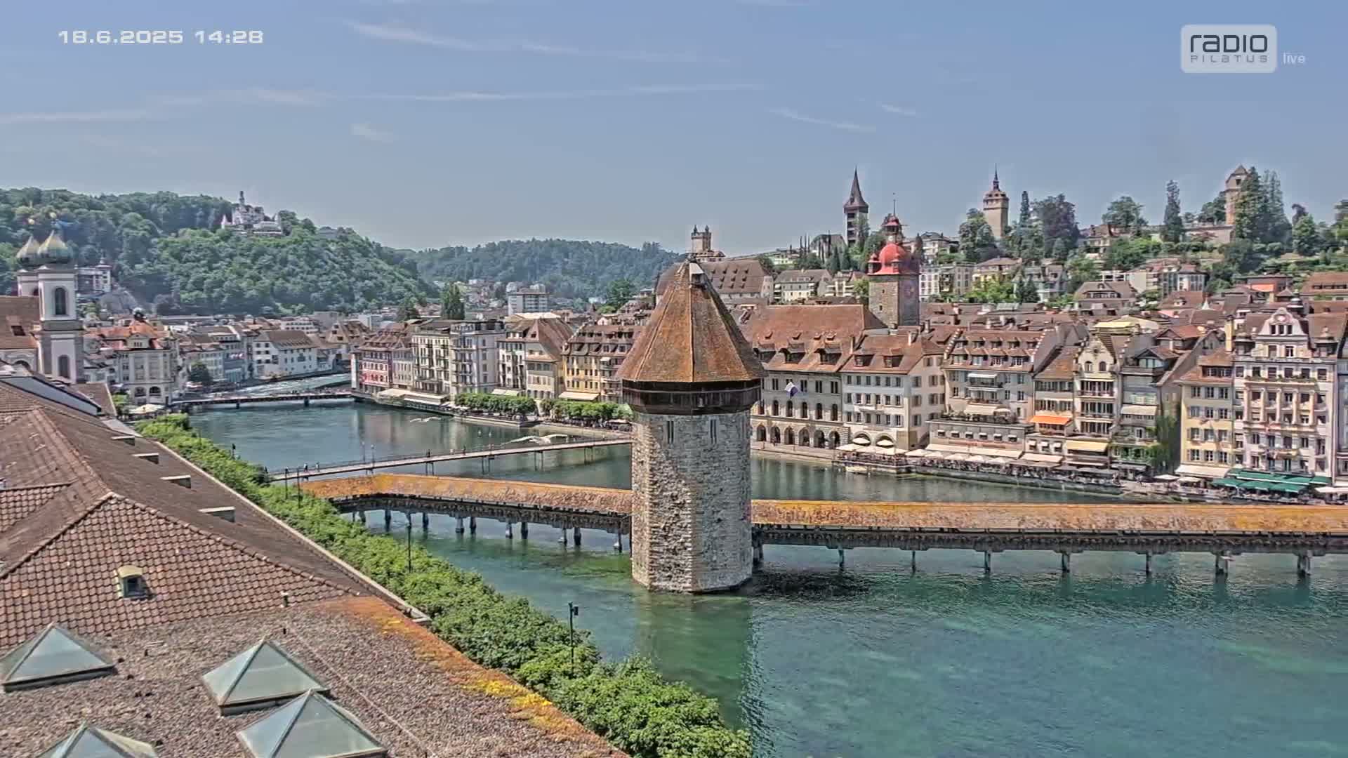 A covered wooden bridge spans a river, with a stone tower in the center, overlooking a town of buildings nestled against a green hill on a sunny day.