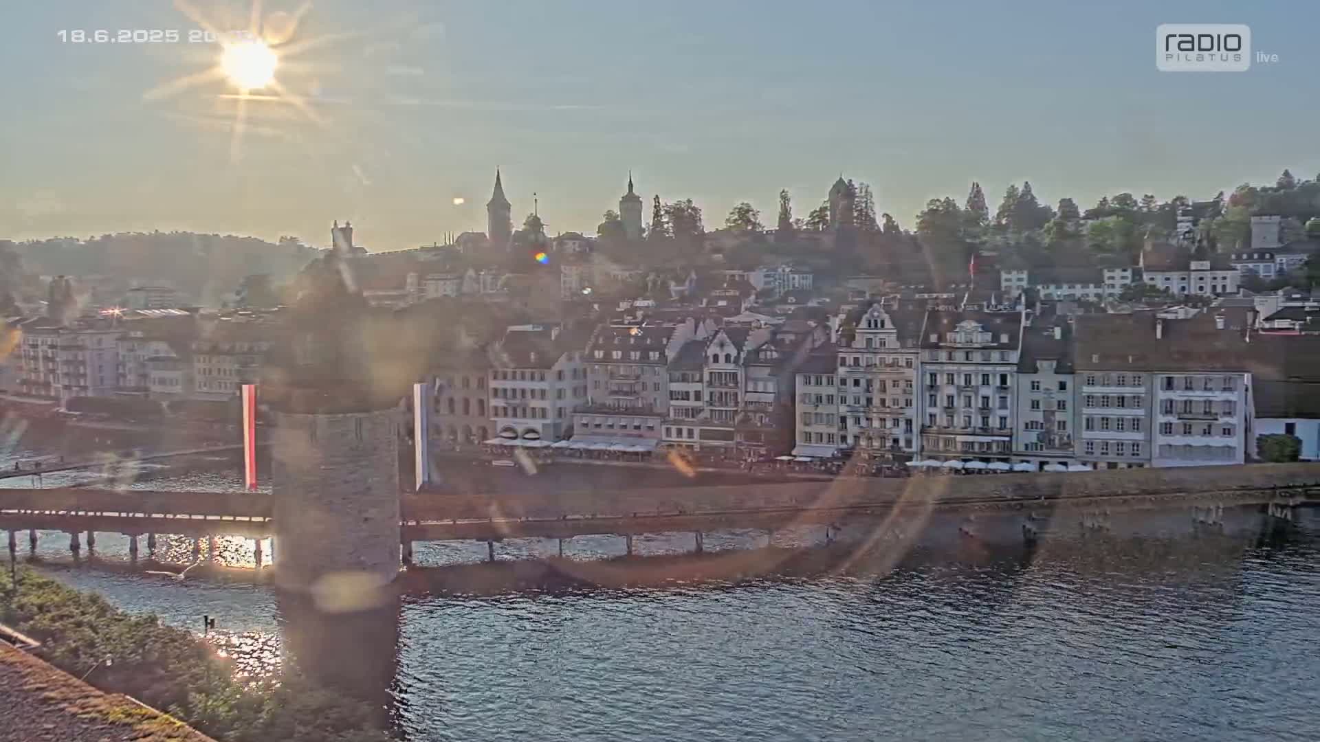 A sunlit view of a European town situated on a river, with a bridge and buildings lining the waterfront under a mostly clear sky.