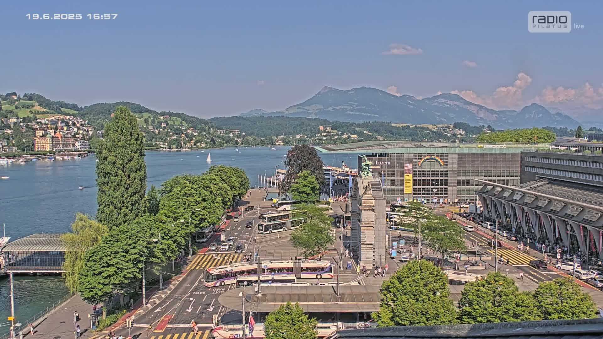 A sunny day overlooking a city's waterfront, featuring buses, cars, and buildings near a lake with mountains in the background.