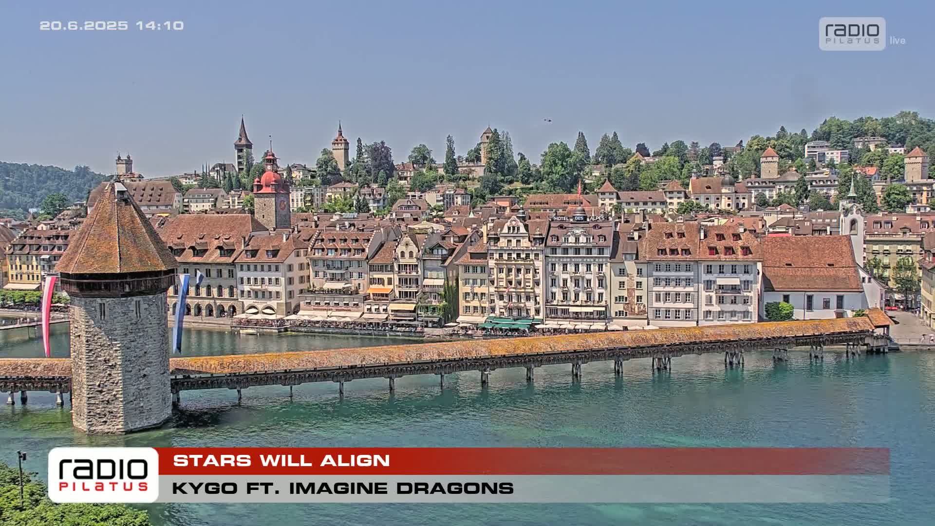 A covered wooden bridge spans a body of water in front of a town with many buildings under a clear blue sky.