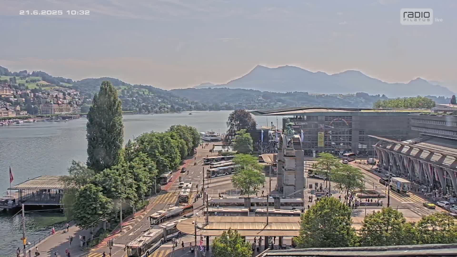 A sunny day overlooking a lake and city with mountains in the background, showing buses, trams, and a large building near a waterfront.