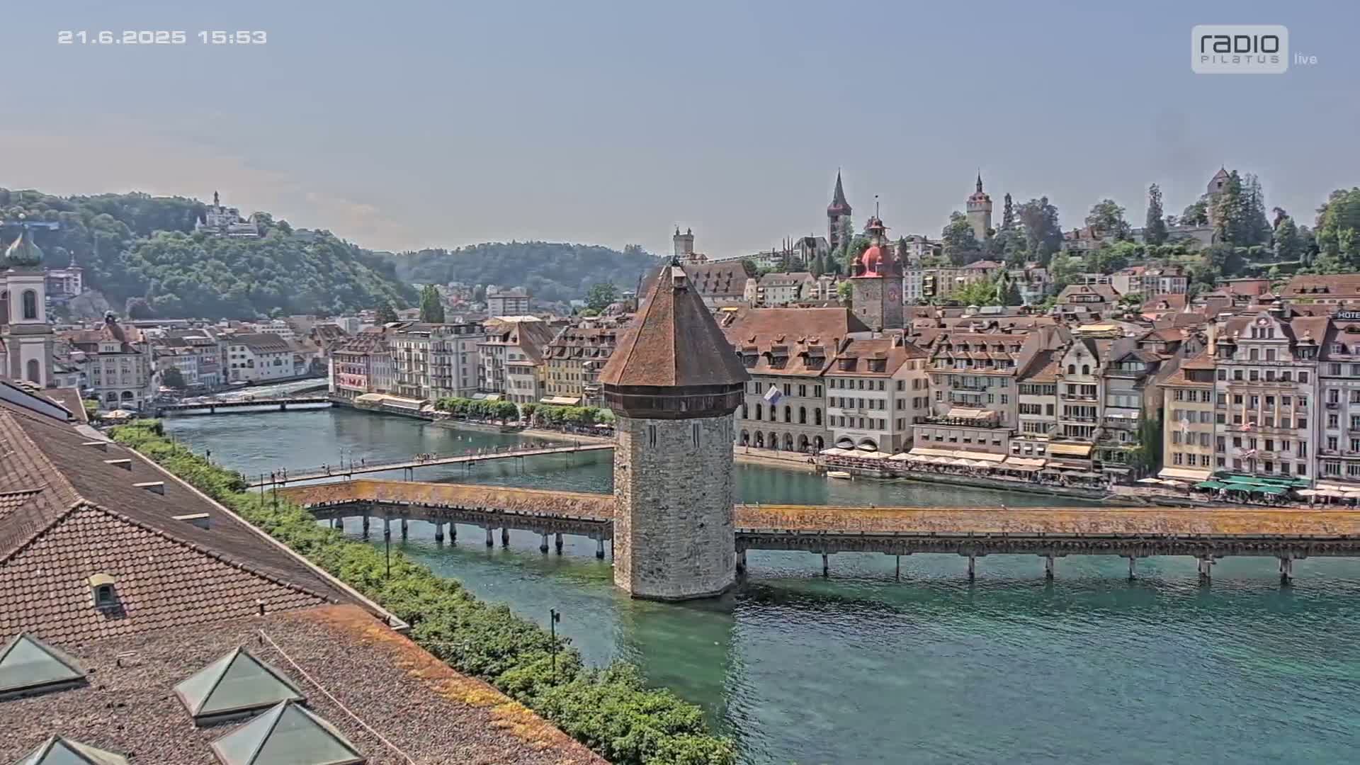 A stone tower stands in the middle of a long wooden bridge spanning a river, with many buildings lining the riverbanks under a clear, sunny sky.