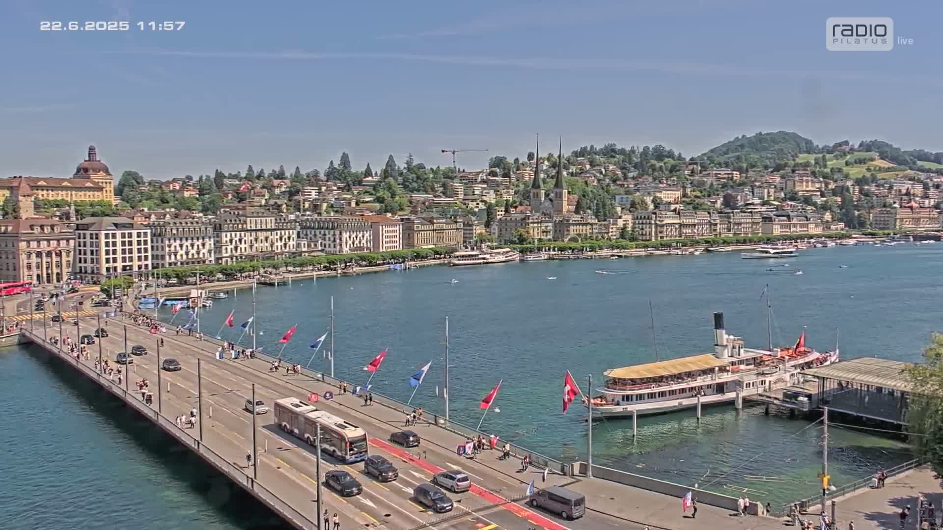 A bridge with traffic and pedestrians spans a body of water, with a city skyline and a paddle steamer docked on the opposite shore under sunny skies.