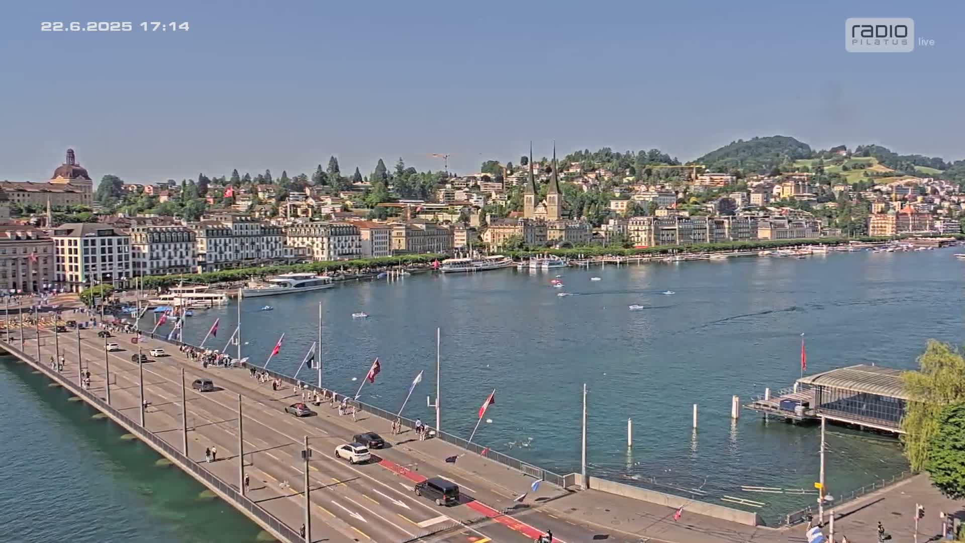 A bridge with cars and pedestrians spans a calm lake, overlooking a city of buildings nestled against a hillside under a clear sky.