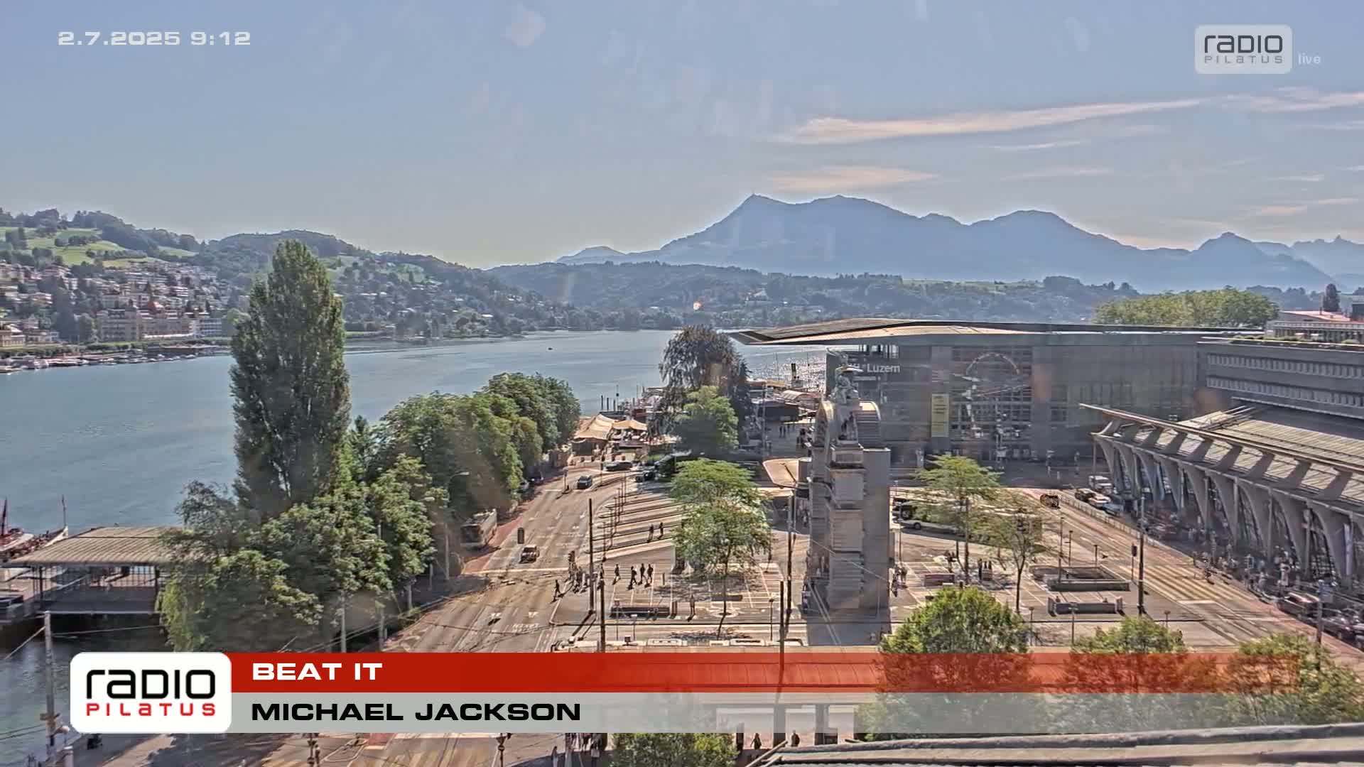 A sunny cityscape overlooks a calm lake and distant mountains, with a city square and buildings in the foreground.