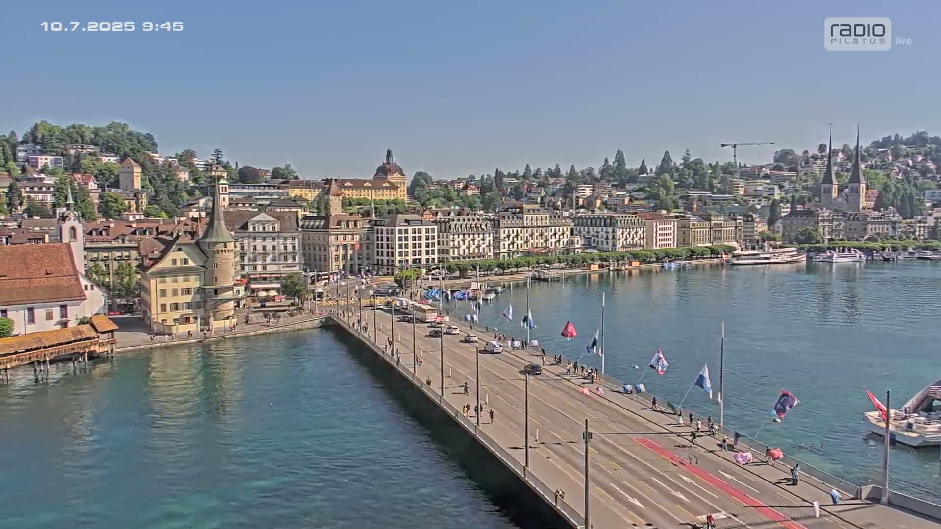 A bridge spans a body of calm, blue water, leading to a town of European-style buildings on a sunny day.