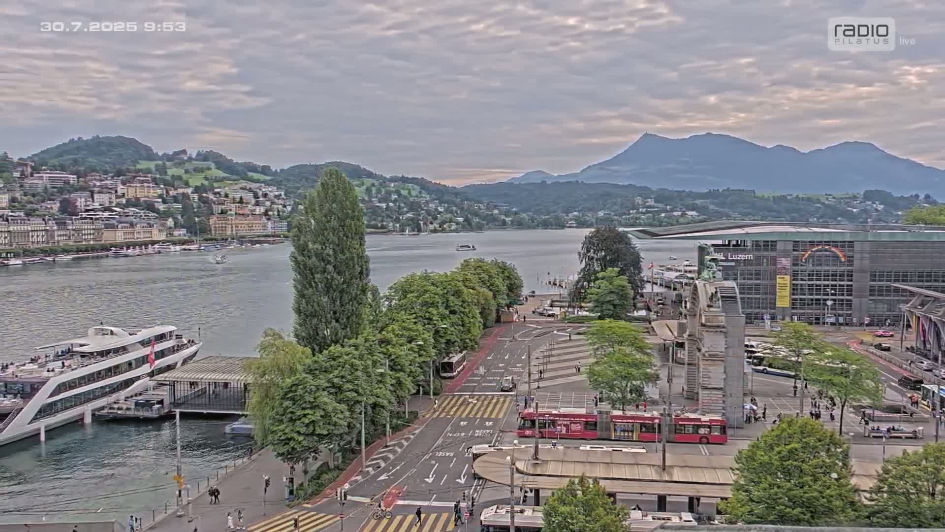A large body of water with a city and mountains in the background under an overcast sky, showing a cruise ship docked near a roadway with buses and pedestrians.