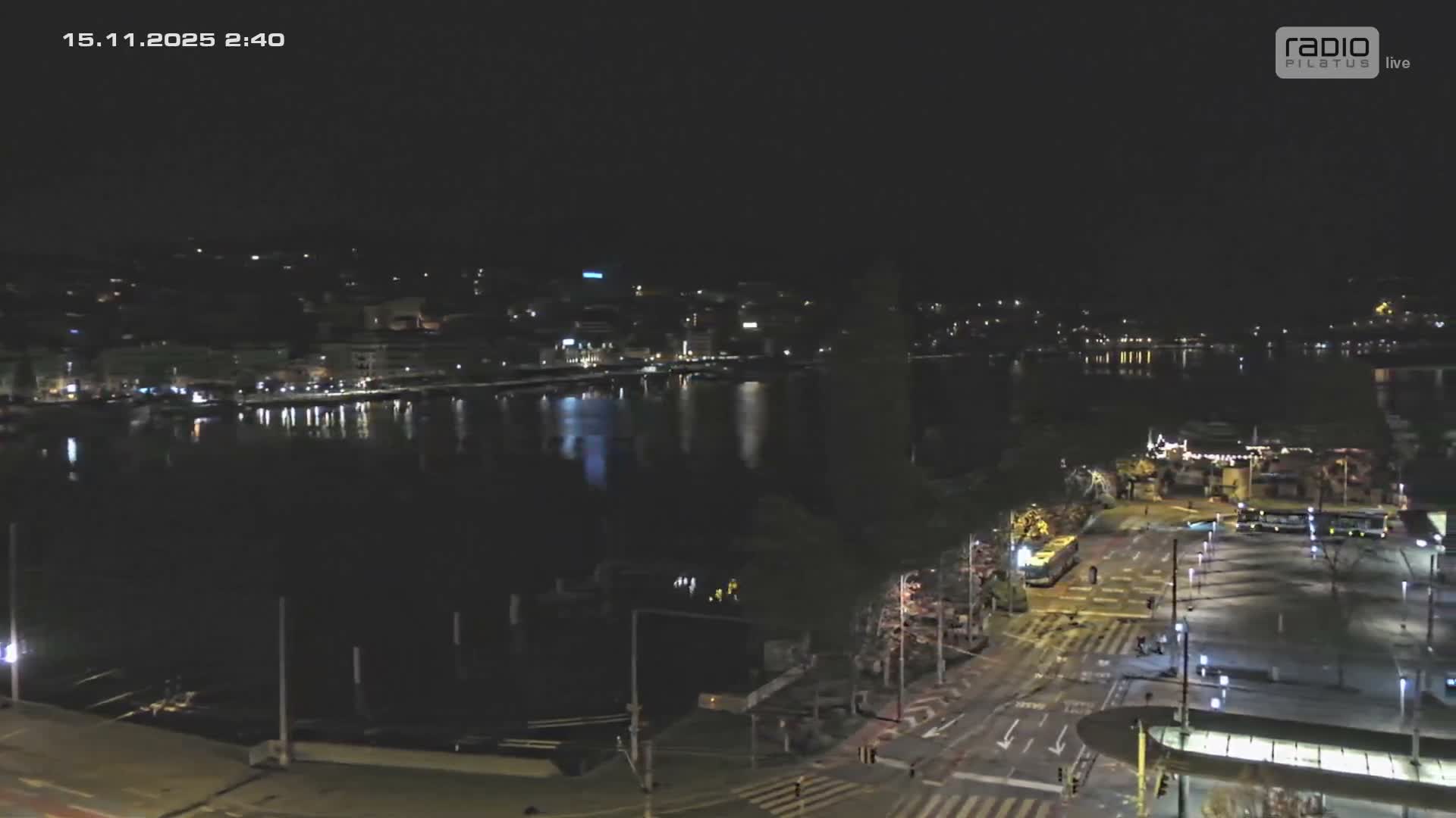 A clear night view captures a city waterfront with lights reflecting on calm water, an illuminated street featuring buses and pedestrians in the foreground, and a distant urban skyline.