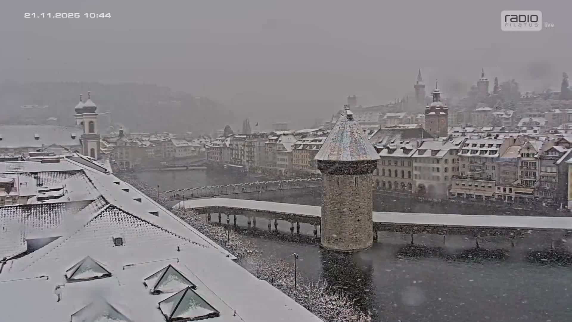 A snow-covered city is depicted under a heavy snowfall, featuring a wide river with a prominent stone tower and bridges, surrounded by buildings with white roofs and distant, hazy hills under an overcast grey sky.