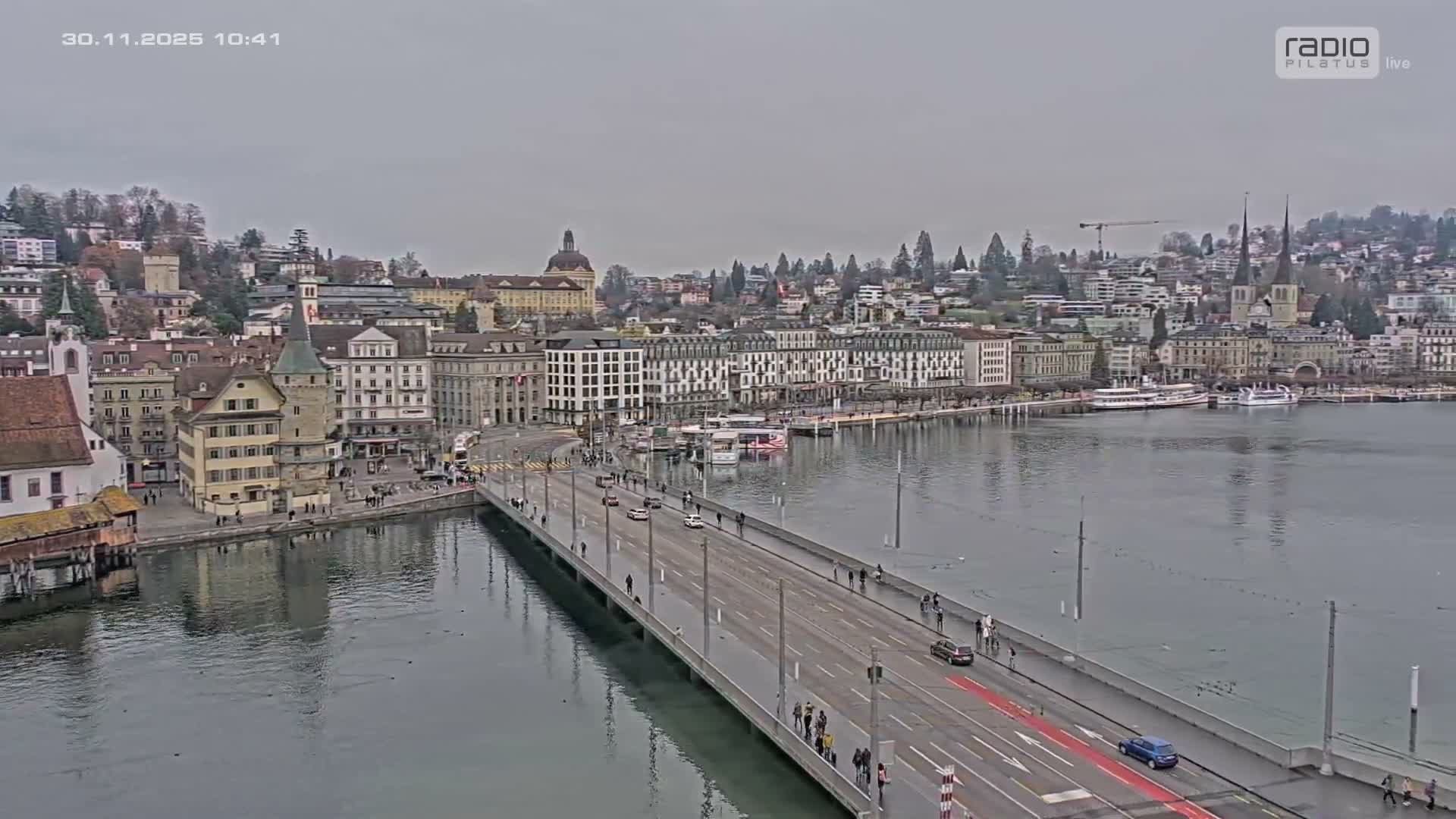 A bustling cityscape on an overcast day features a wide bridge crossing a body of water, bordered by a dense array of historical and modern buildings, many with distinctive spires, and boats docked along the shores, with the city climbing hills in the background.