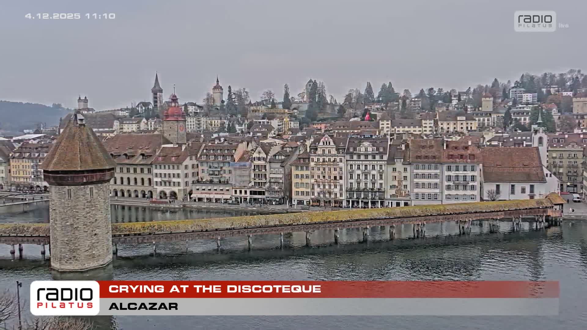A historic European city is viewed on an overcast day, featuring a prominent covered wooden bridge and stone tower spanning a calm river, with numerous multi-story buildings and spired churches rising along the waterfront and up a distant hillside.