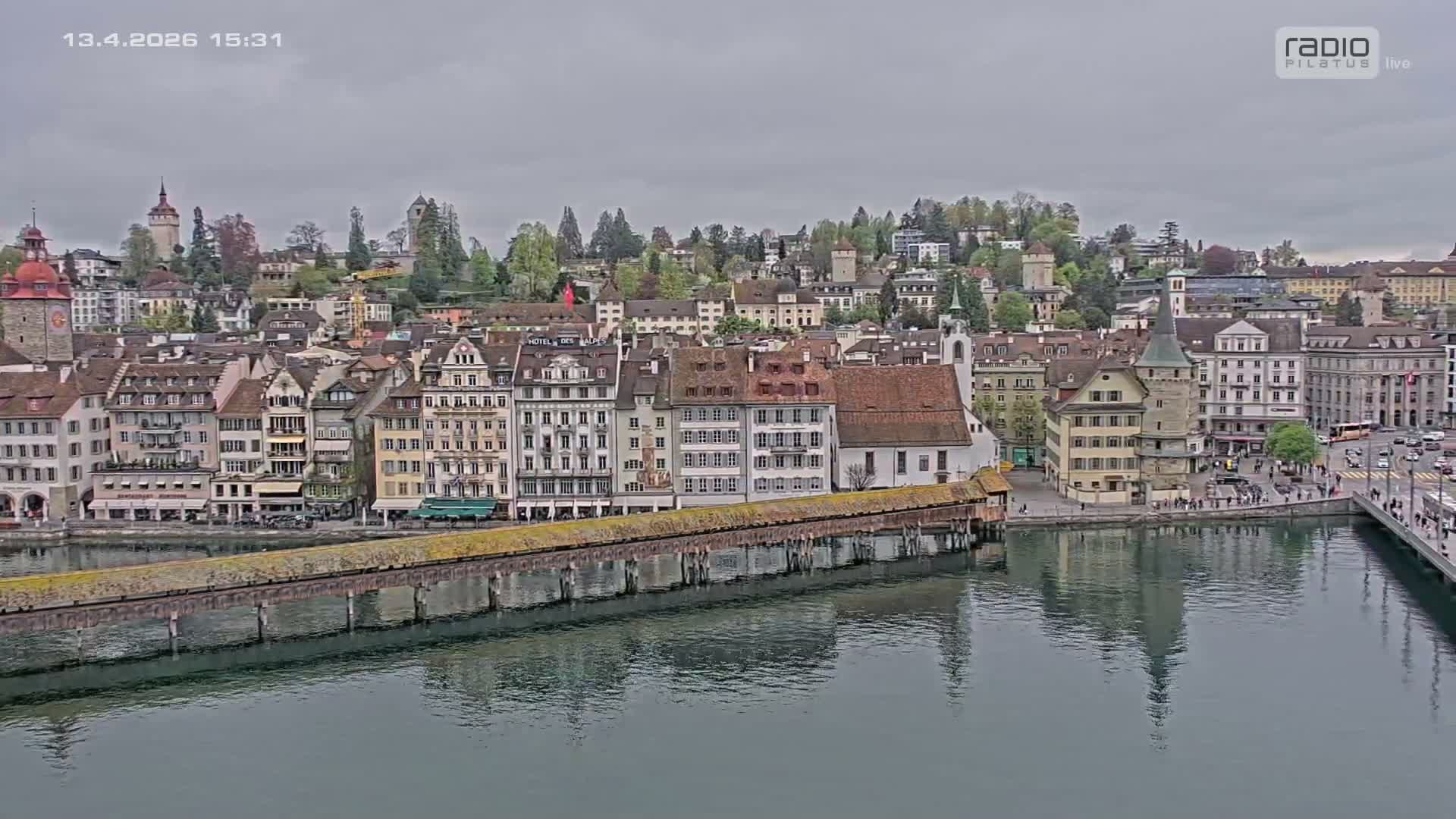 A historic European city is viewed on an overcast day, featuring a prominent covered wooden bridge and stone tower spanning a calm river, with numerous multi-story buildings and spired churches rising along the waterfront and up a distant hillside.