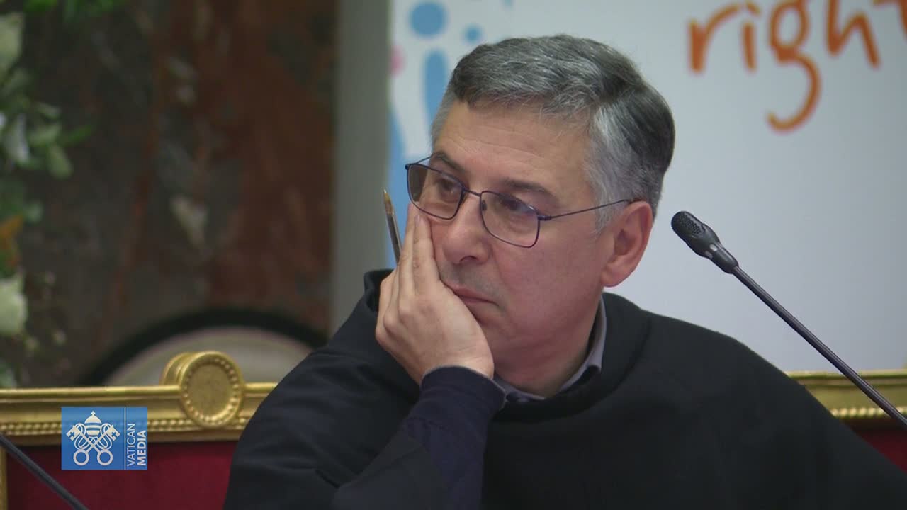 A man with graying hair and glasses sits at a table, his hand resting on his chin, appearing pensive.