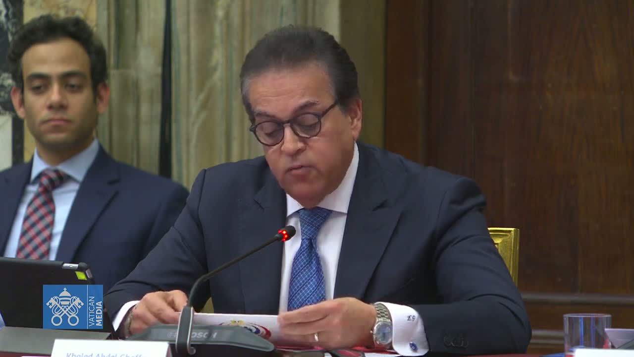 A man in a suit and glasses sits at a table, looking down at papers, while another man in a suit sits behind him.