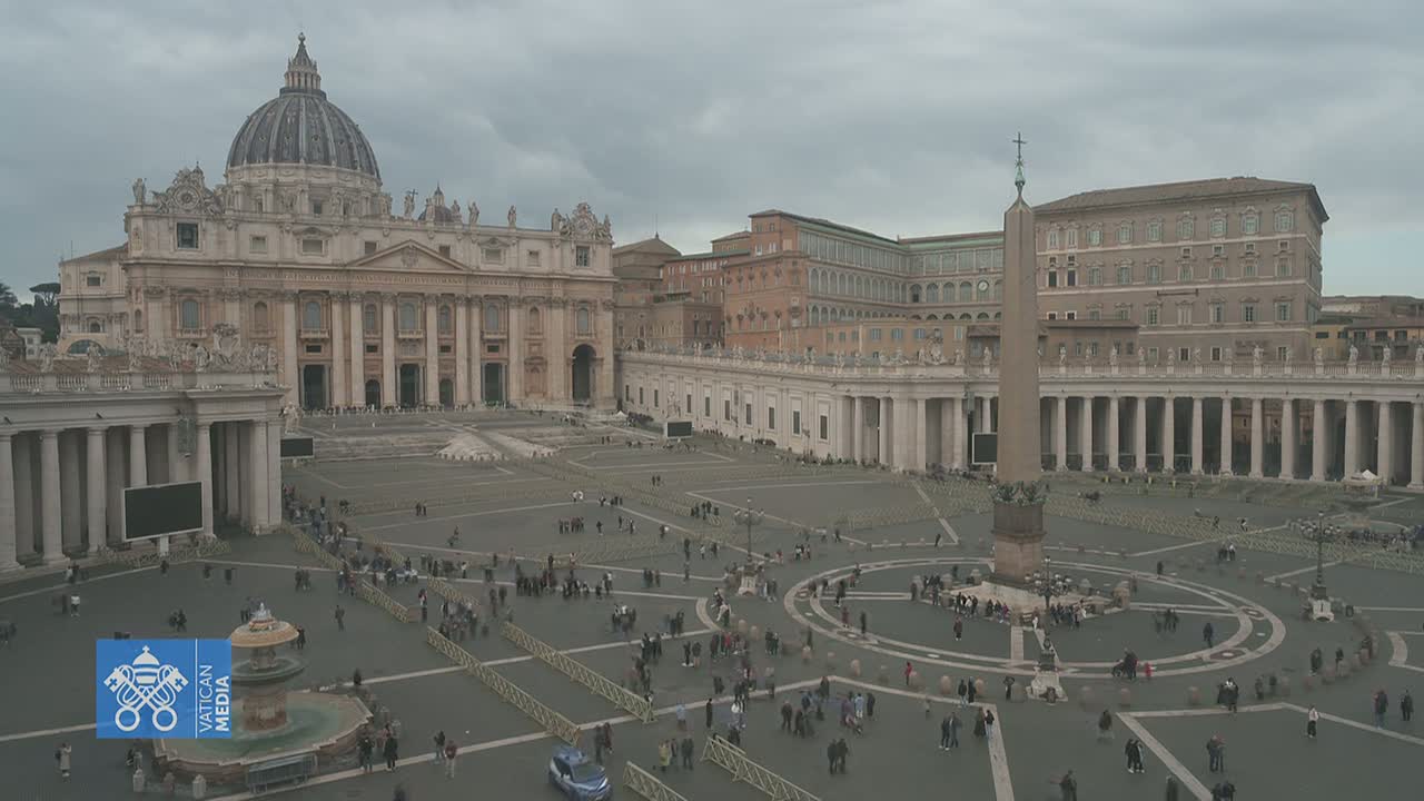 St. Peter's Square in Vatican City, under a cloudy sky, is sparsely populated with people walking and standing around the large, paved plaza.