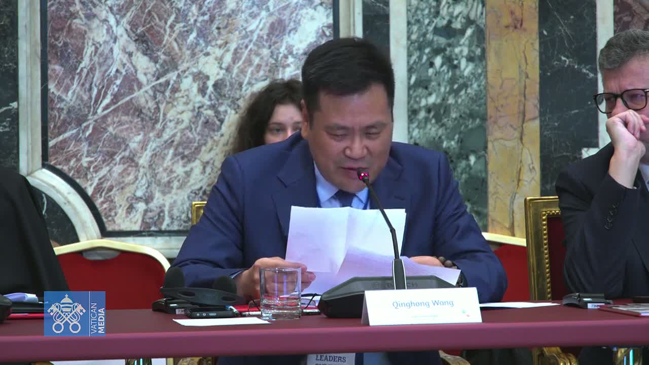 A man in a dark blue suit sits at a table, reading papers while speaking into a microphone, with another man in a suit seated to his right.