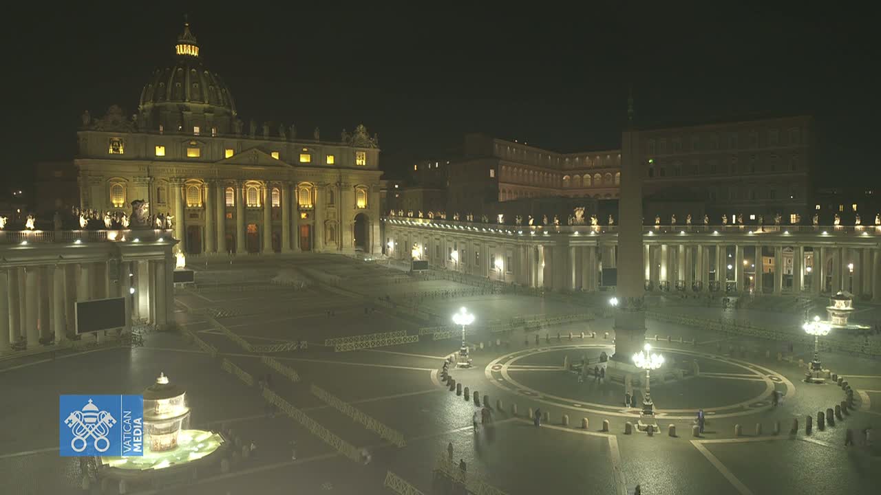 St. Peter's Basilica and Square are illuminated at night, with a few people visible in the vast, mostly empty space.