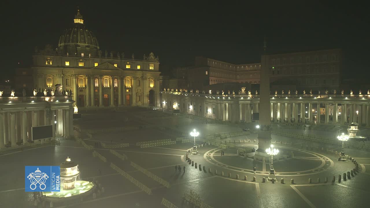 St. Peter's Basilica and St. Peter's Square are illuminated at night, sparsely populated, with barriers and lampposts visible throughout the square.