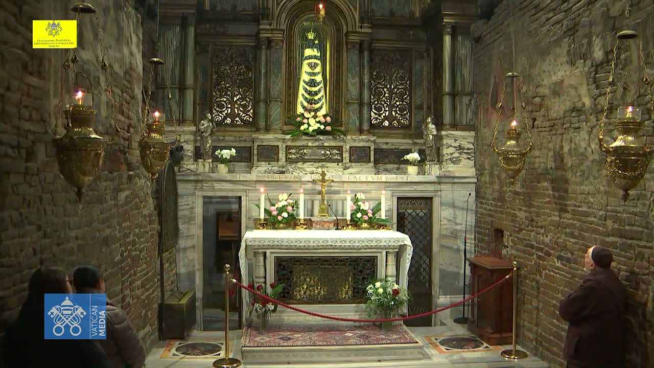 An altar with candles and flowers sits in a dimly lit stone chapel, where a man in brown robes stands near a brick wall and two people are observing.