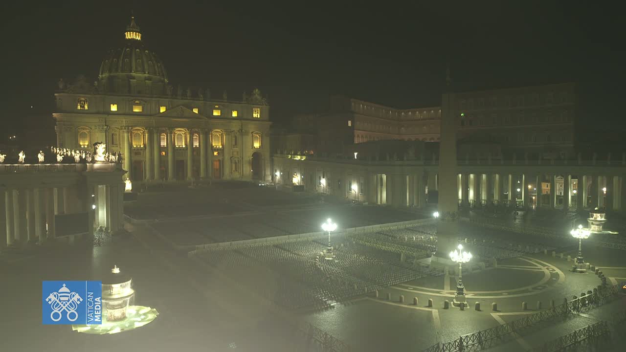St. Peter's Basilica and Square are illuminated at night, showing a mostly empty plaza with rows of chairs set up under a hazy sky.
