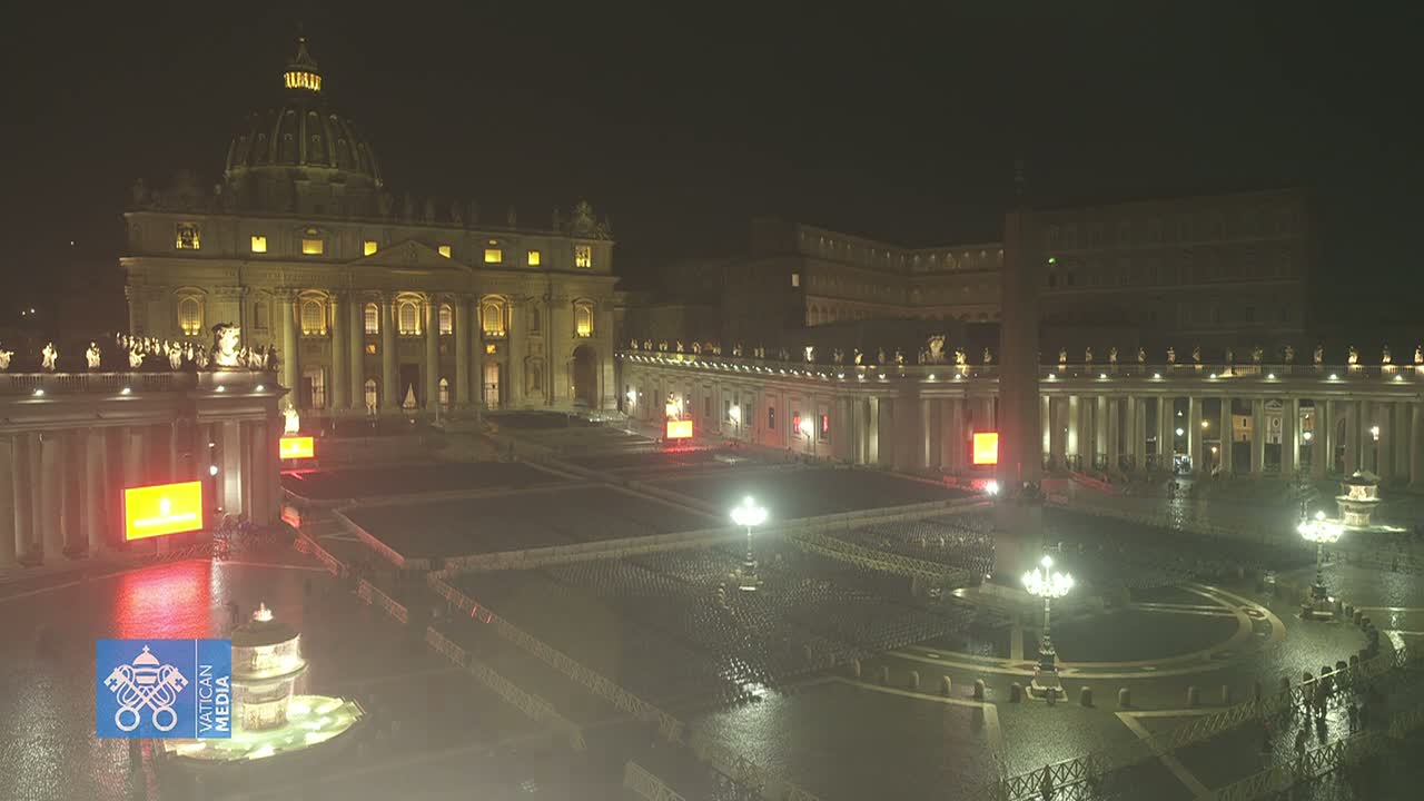 A nighttime, rain-slicked St. Peter's Square in Vatican City shows St. Peter's Basilica and surrounding buildings lit up, with seating arranged in the square.