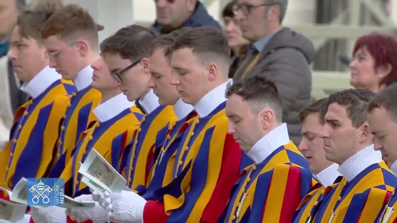 A row of men in blue, yellow, and red uniforms stand outdoors, heads bowed, holding pamphlets.