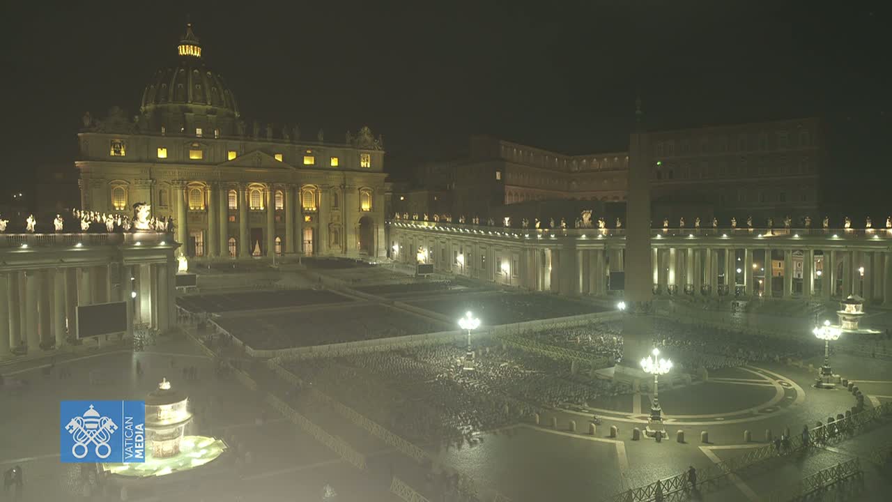 A nighttime, aerial view of St. Peter's Basilica and Square in Vatican City, showing a large gathering of people illuminated by lights.