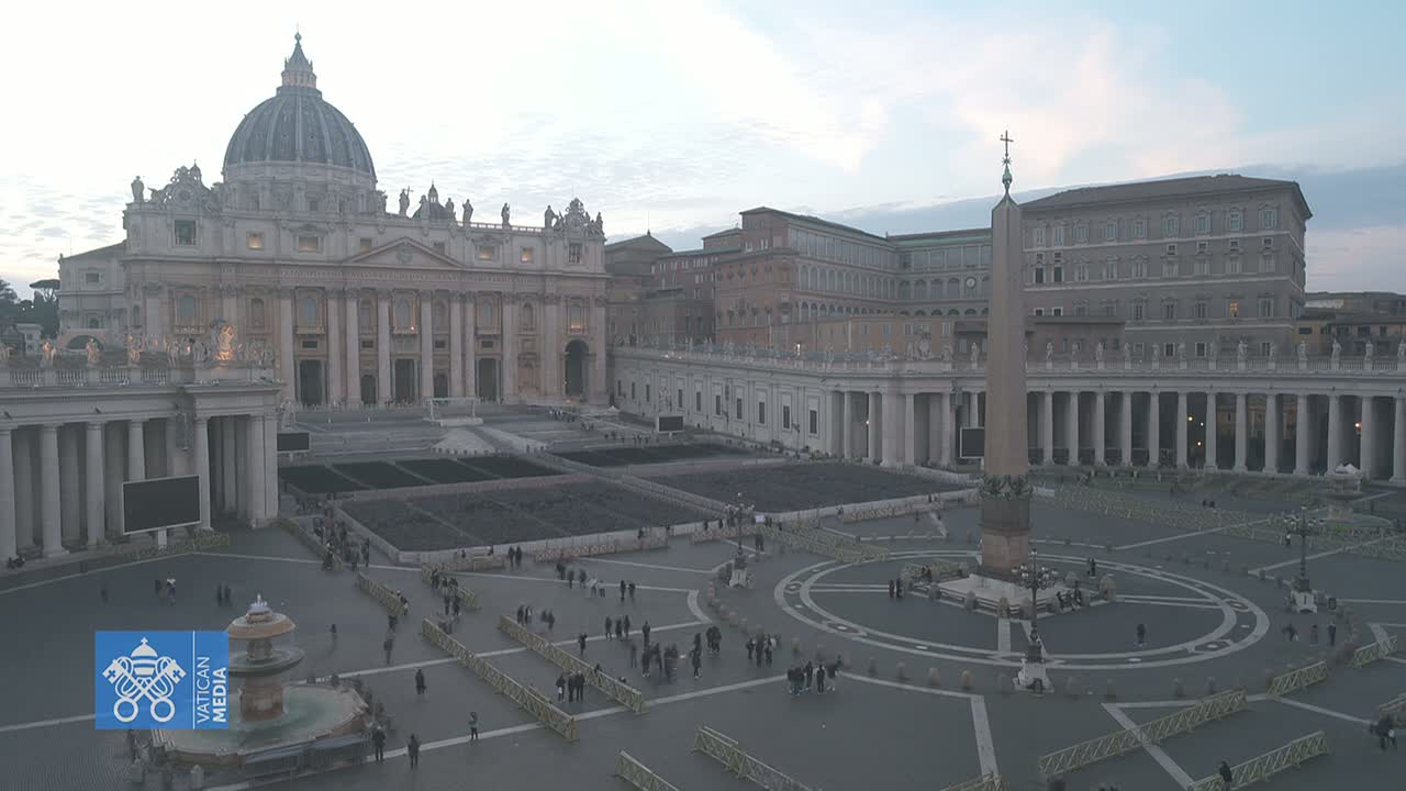 St. Peter's Square in Vatican City, under a dusky sky, shows a large expanse of paved area with a few people and barriers, centered around an obelisk, with St. Peter's Basilica and other Vatican buildings in the background.