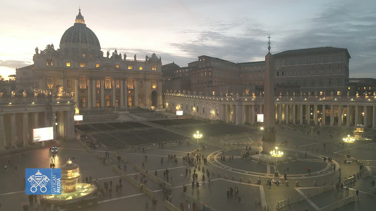 St. Peter's Basilica and St. Peter's Square at dusk, with a few people scattered throughout the square under a cloudy sky.