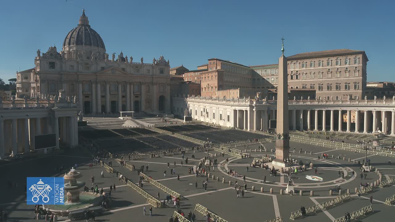 St. Peter's Square in Vatican City on a sunny day shows many people sparsely scattered across the large plaza, with rows of empty chairs set up, and St. Peter's Basilica in the background.