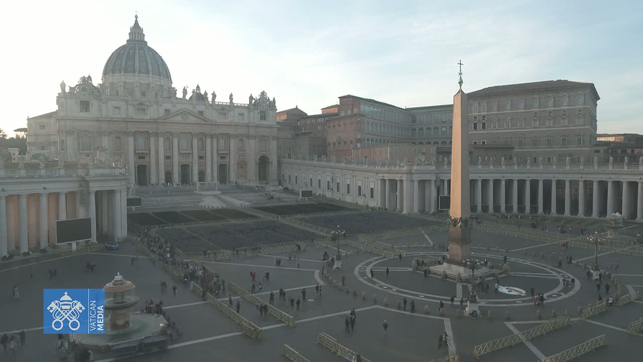 A high-angle, long shot shows St. Peter's Basilica and Square in Vatican City on a hazy, overcast day, with a few people scattered throughout the large plaza.