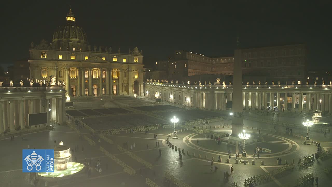A nighttime, illuminated view of St. Peter's Square and Basilica in Vatican City, with a few people sparsely scattered throughout the vast plaza.