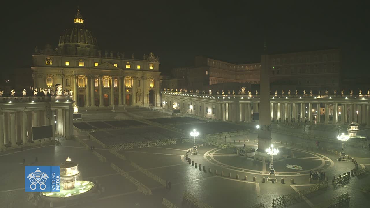 St. Peter's Basilica and Square are illuminated at night, with a few people visible in the vast, mostly empty plaza.