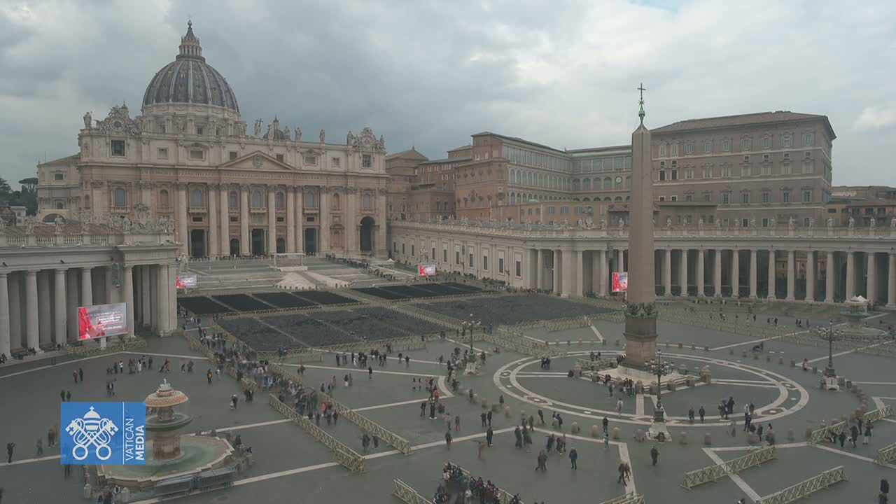 St. Peter's Square in Vatican City, under a cloudy sky, shows a large crowd of people gathered in front of St. Peter's Basilica.