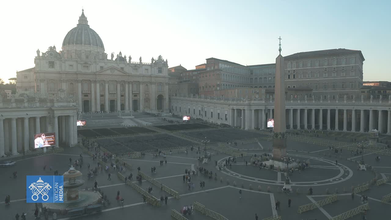 St. Peter's Square in Vatican City, under a pale, dusky sky, shows a large crowd gathered around a central obelisk, with St. Peter's Basilica in the background.